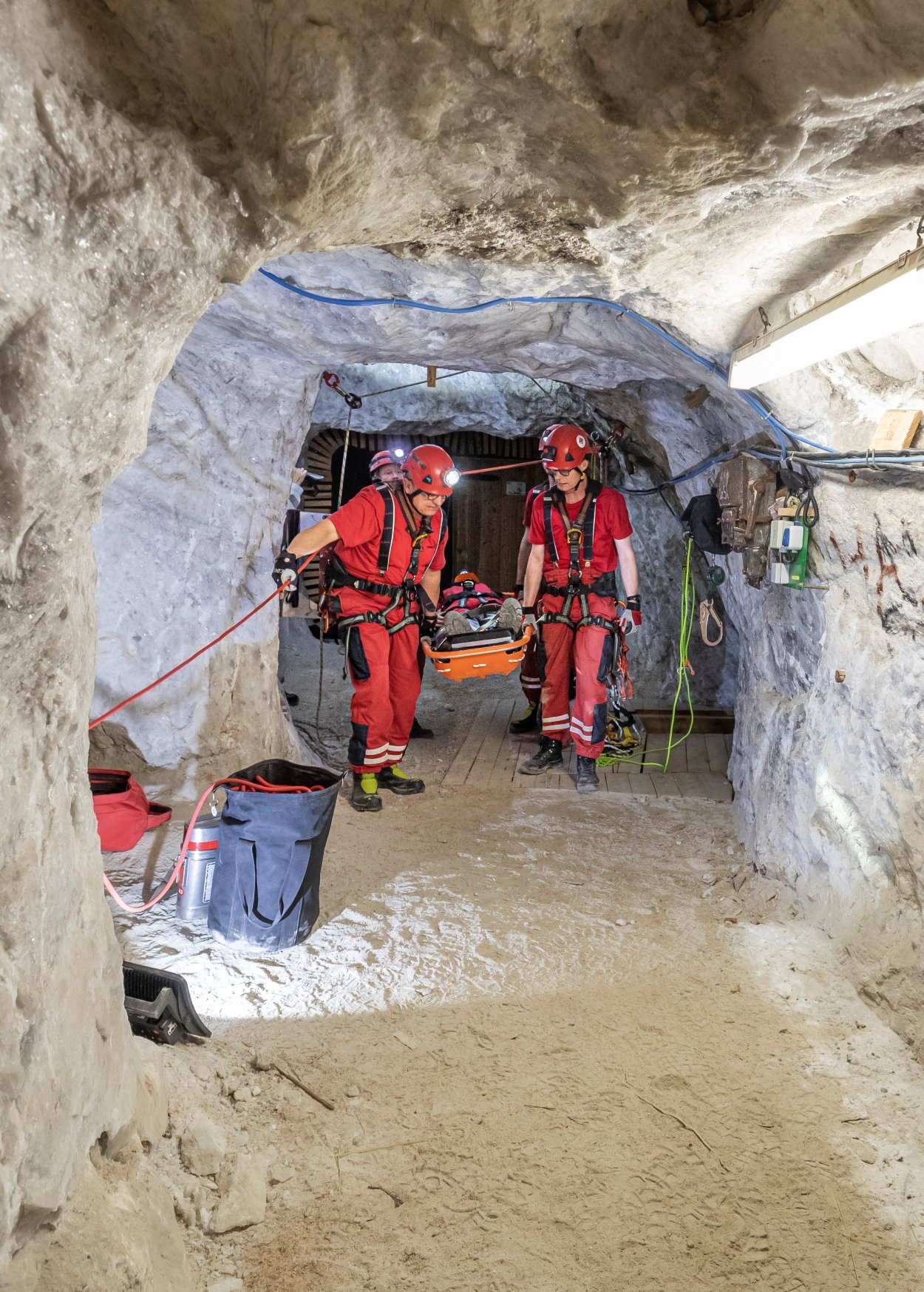 Zwei Männer in roten Arbeitsanzügen gehen durch einen Bergwerkschacht. Zwischen ihnen tragen sie eine Trage mit einem Verletzen.