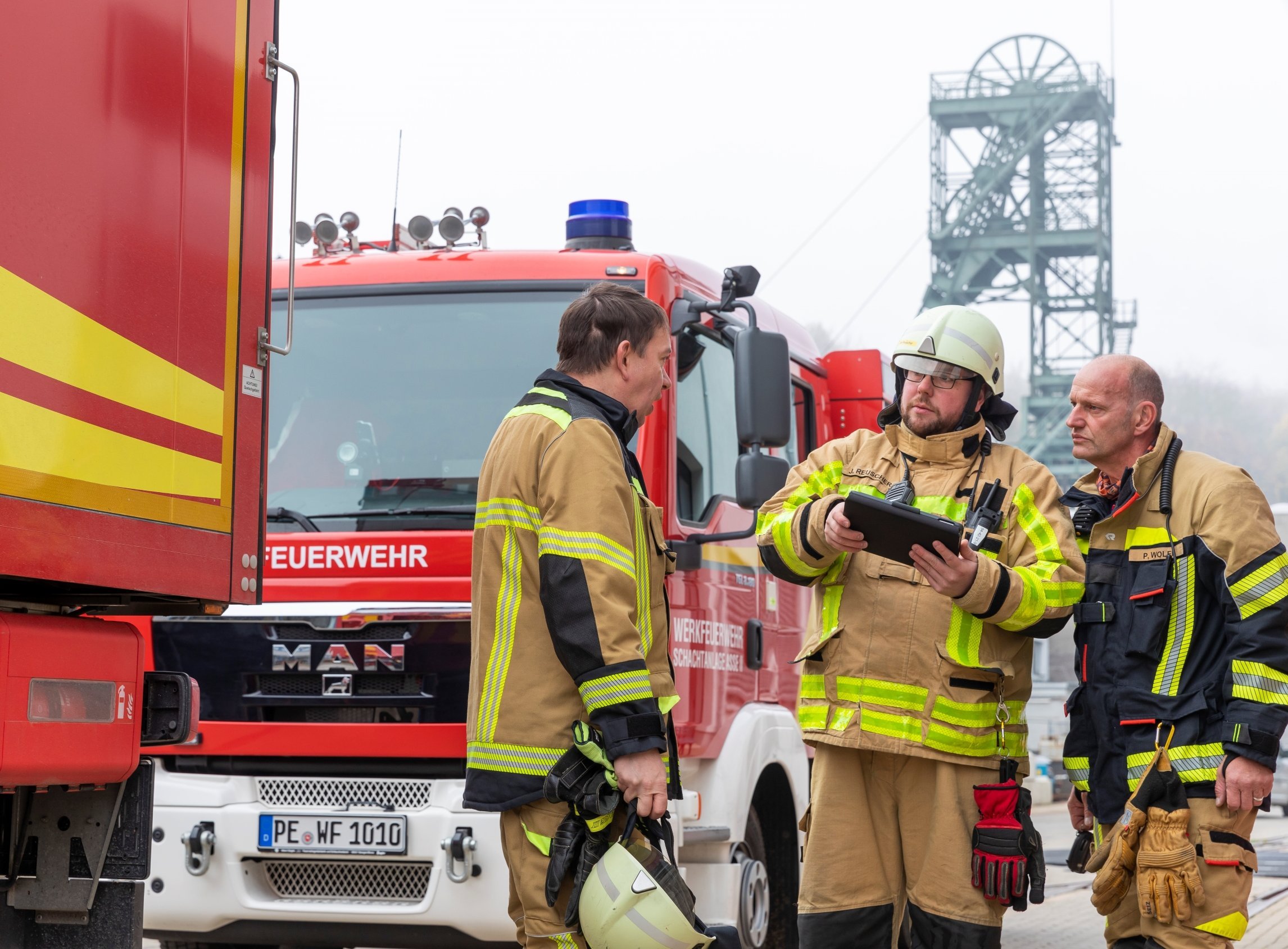 Drei Männer in braunen Feuerwehruniformen stehen vor einem Feuerwehrfahrzeug.