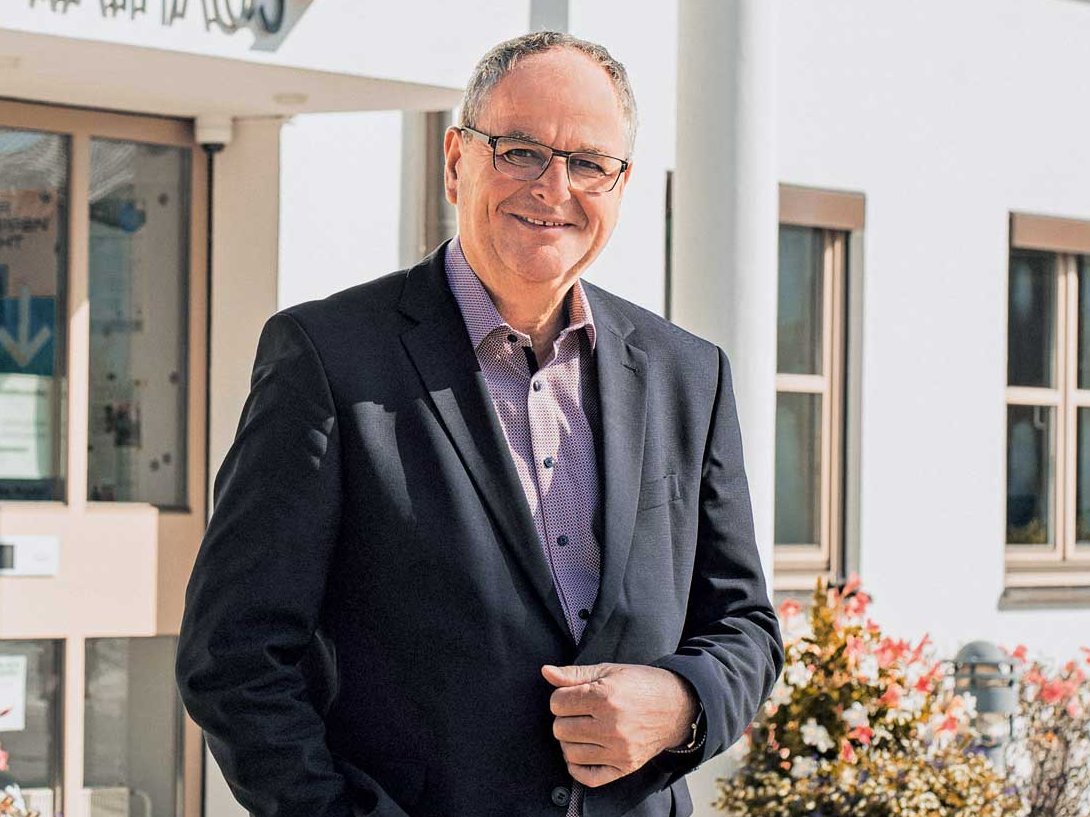 Vor einem Haus steht ein Mann in schwarzem Anzug. Er trägt eine Brille sowie ein fliederfarbenes Hemd.