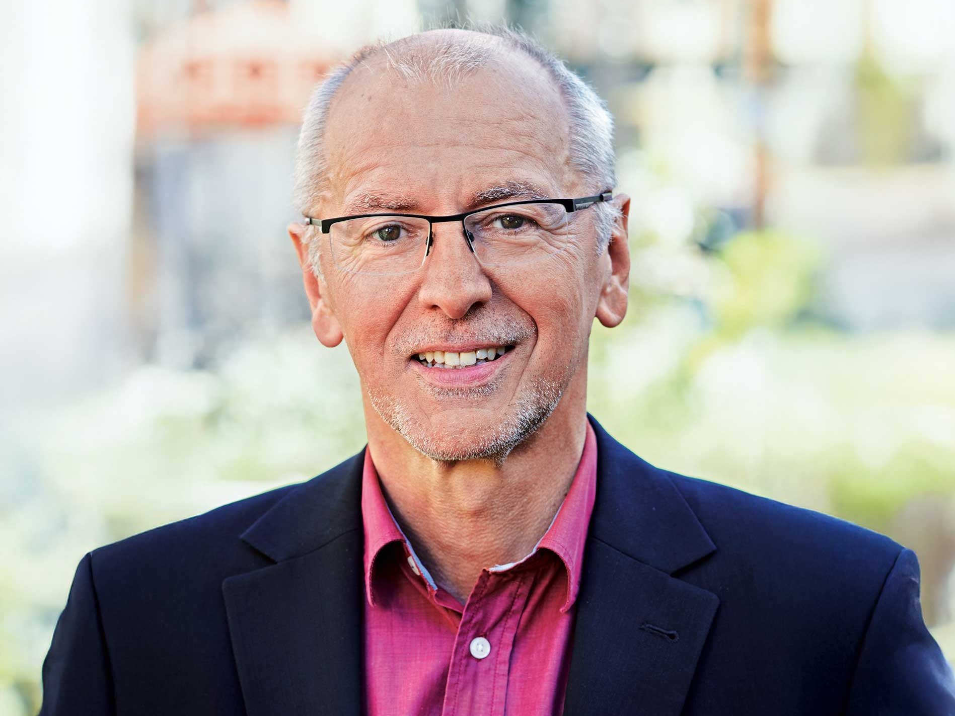 Ein Mann mit grauen Haaren und grauem Bart lächelt in die Kamera. Er trägt eine Brille, ein pinkfarbenes Hemd und ein schwarzes Sakko.