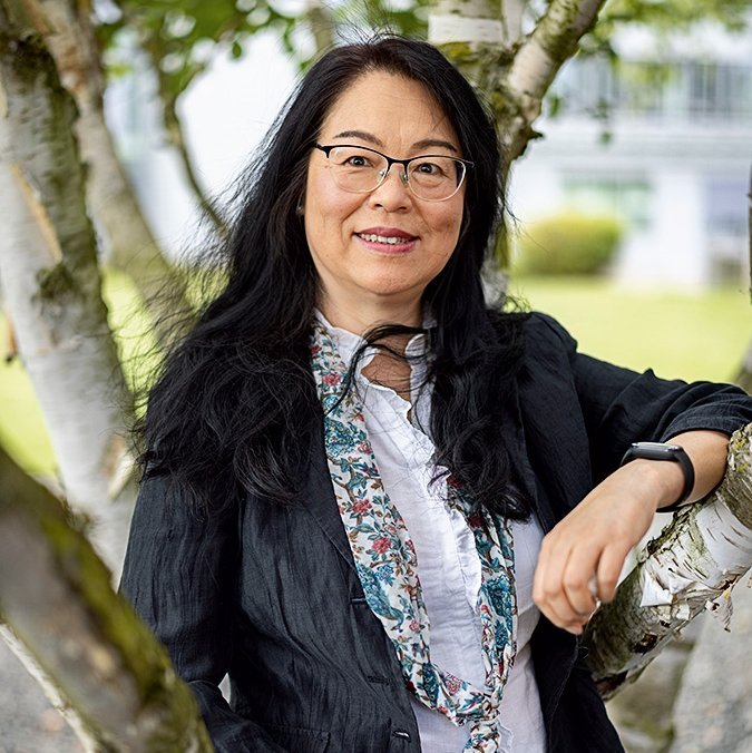 Eine Frau mit schwarzen langen Haaren und Brille lehnt an einem Baum. Sie trägt eine bunte Bluse unter einem dunklen Blazer.