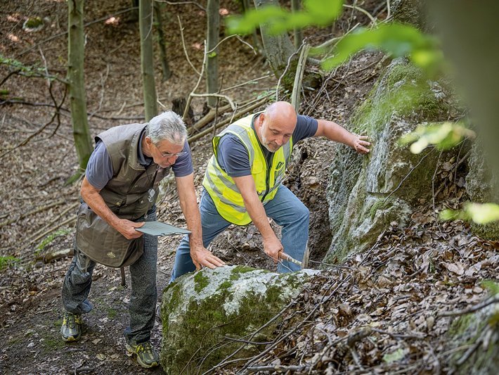 Zwei Männer stehen in einem Wald und inspizieren vor ihnen befindliches Gestein. Einer der Männer hält ein Klemmbrett in der Hand, der andere hält einen Hammer. 