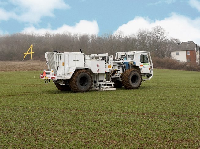 Auf einem Feld steht ein großes weißes Fahrzeug. Im Hintergrund ist ein Wald zu sehen vor blauem Himmel.