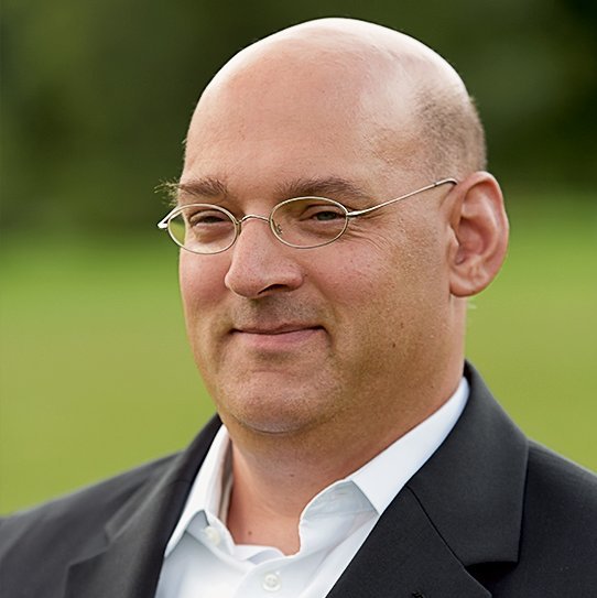 Poträtfoto: Ein Mann mit Glatze und Brille lächelt seitlich in die Kamera. Er trägt ein weißes Hemd unter einem schwarzen Sakko.