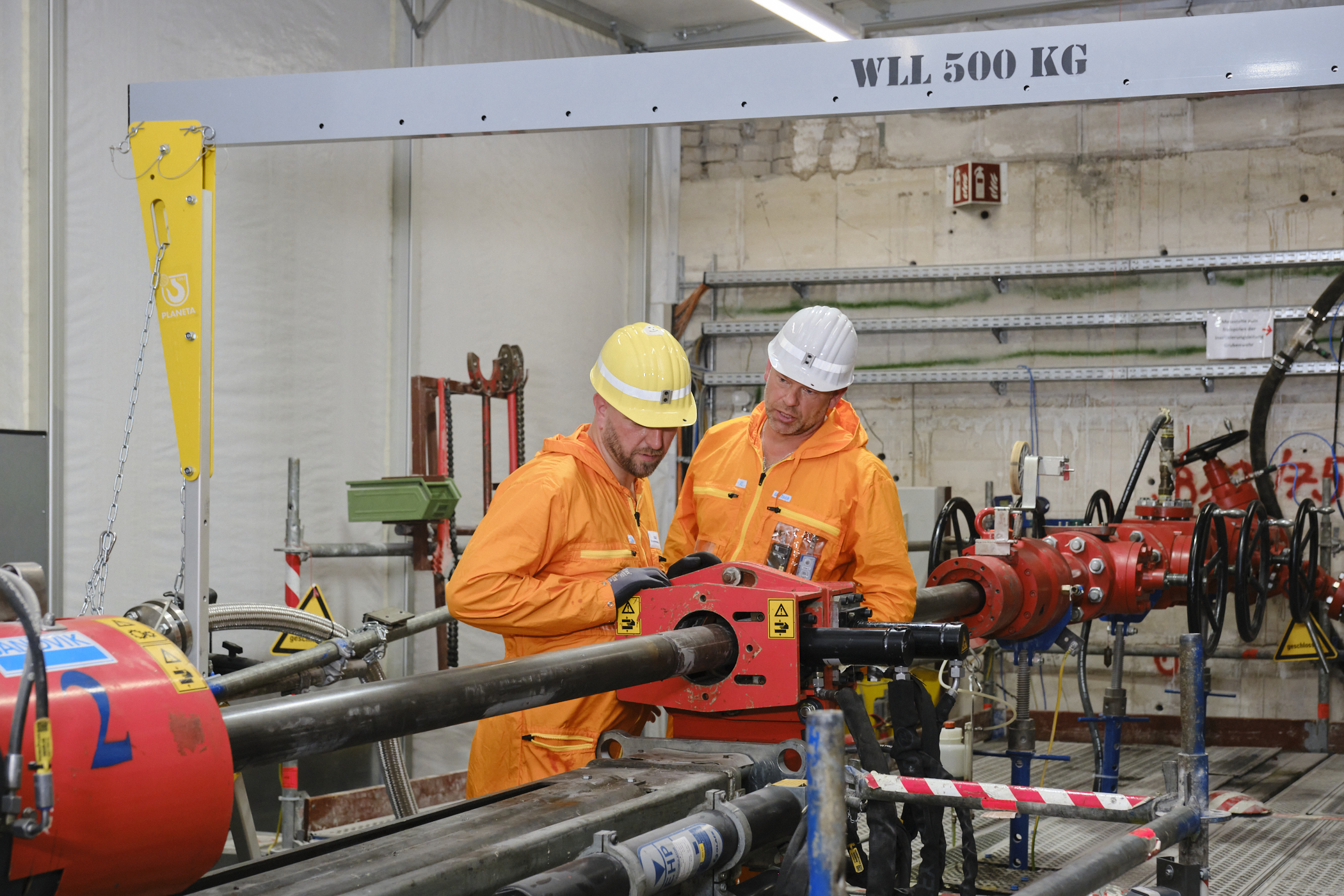 Zwei Männer in orangefarbenen Arbeitsanzügen und Schutzhelmen stehen vor einem vertikalen Bohrstrang.