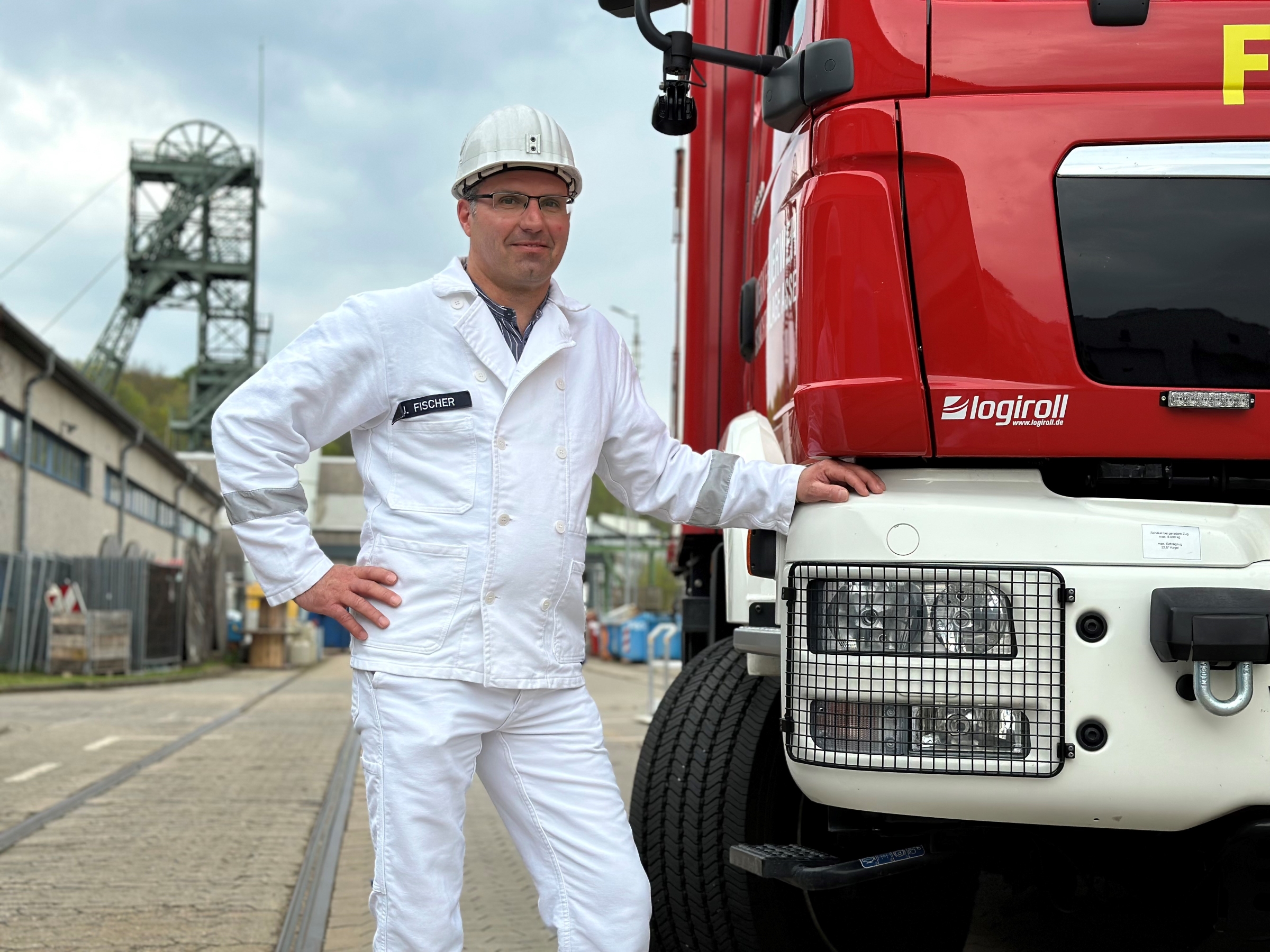 Ein Mann in weißem Anzug und weißem Helm lehnt mit der Hand an eine Feuerwehrfahrzeug.