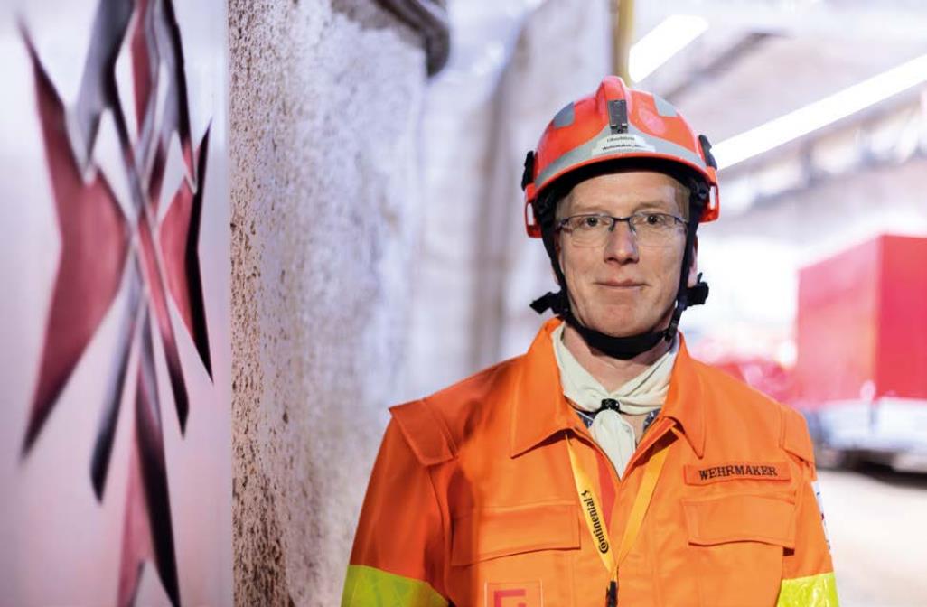 Ein Portrait von einem Mann in orangener Jacke und Helm vor eine Wand unter Tage