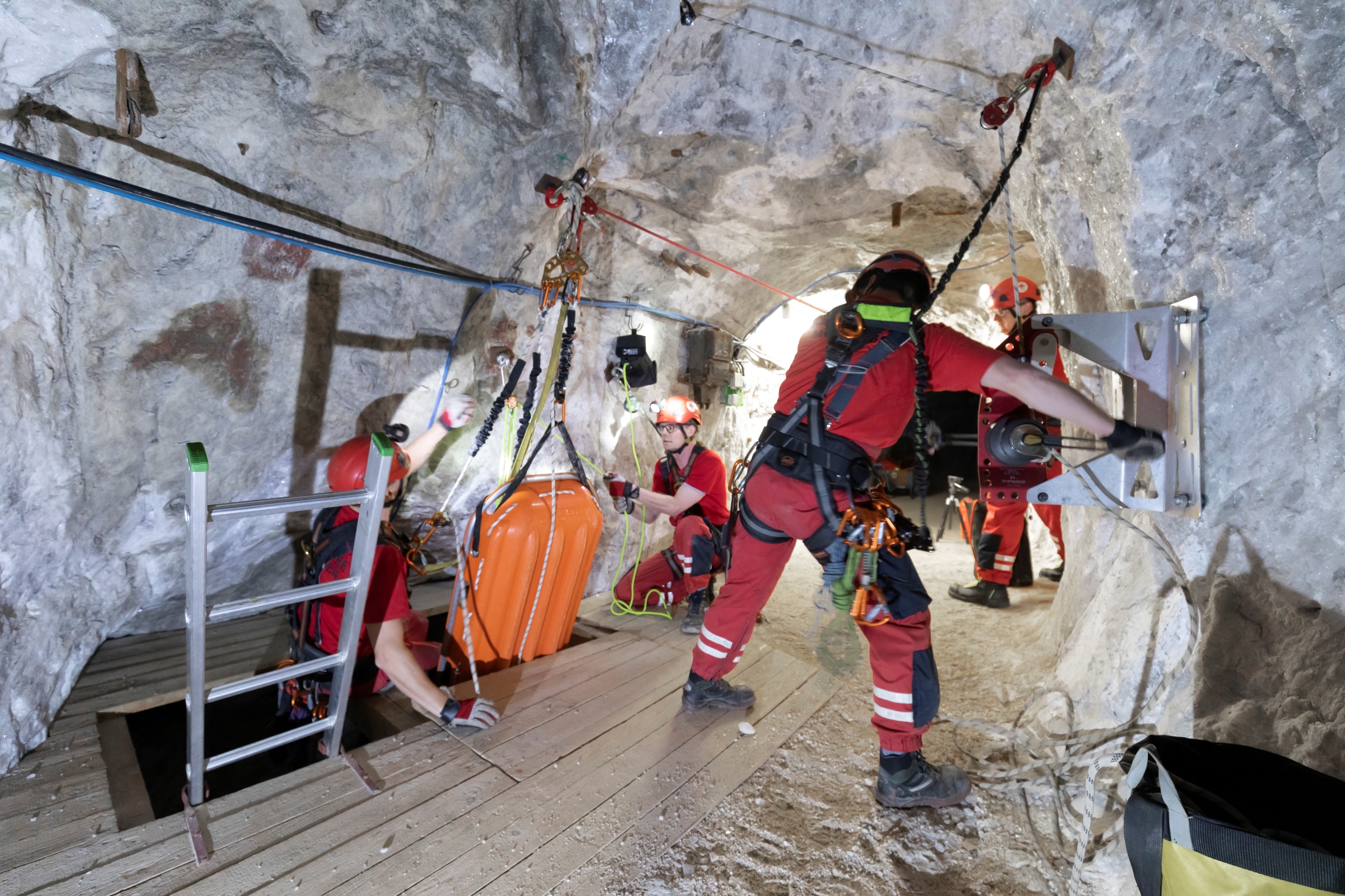 In einem Bergwerkschacht ziehen 4 Männer in roten Arbeitsanzügen eine Trage aus der Tiefe.