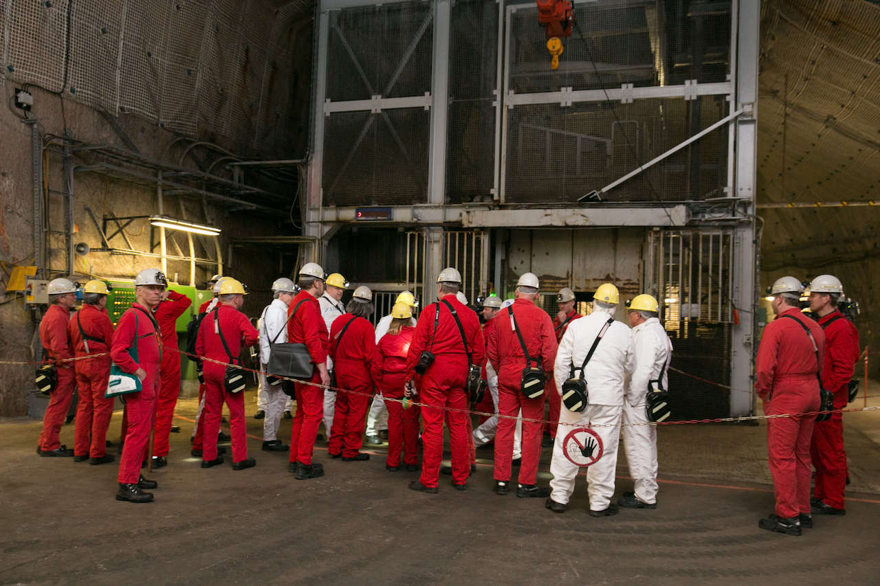 Eine Gruppe Personen in roten Overalls vor einem Aufzug des Bergwerks