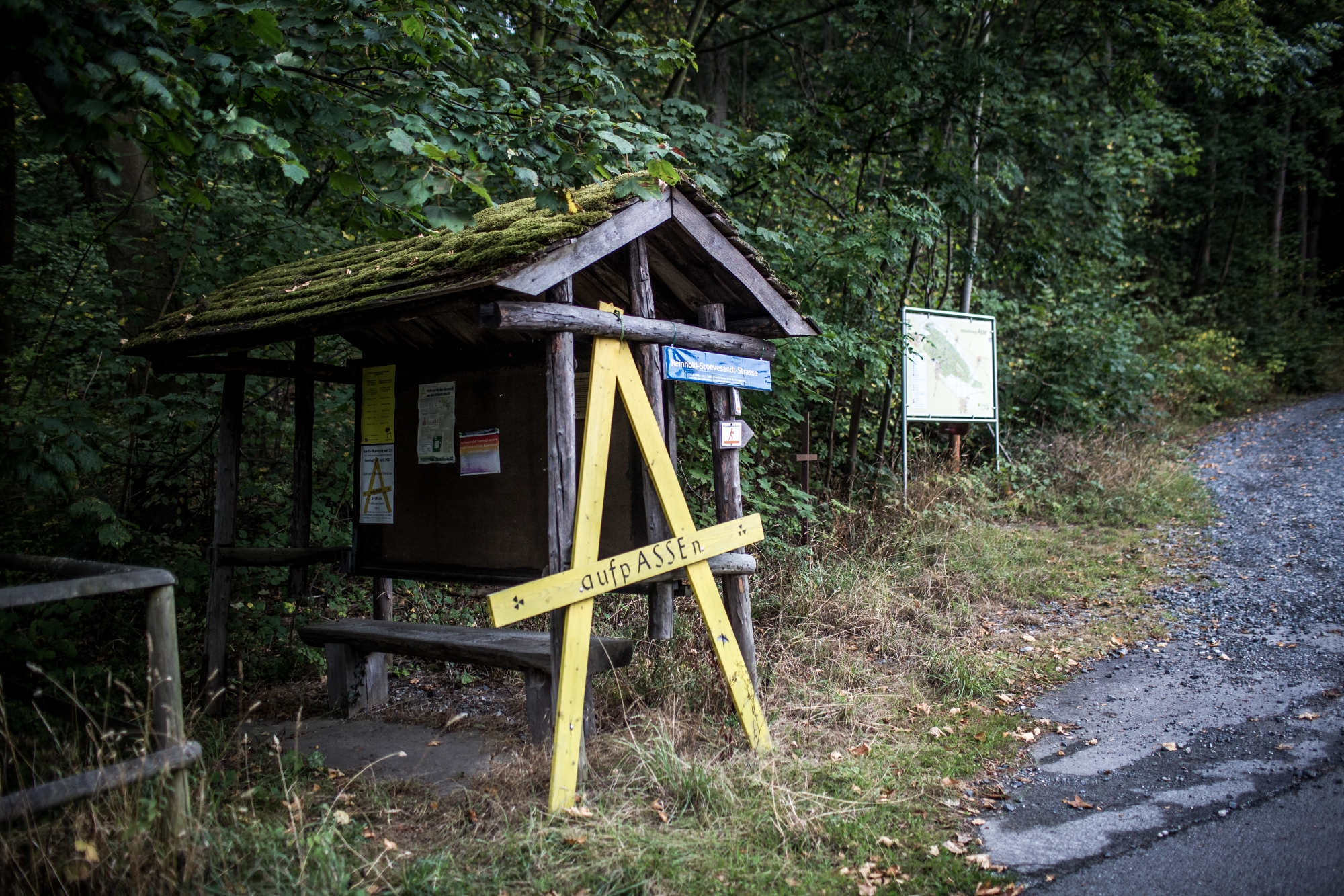 Ein großes gelbes Holzschild in Form des Buchstaben "A" lehnt an einem überdachten Infostand aus Holz an einem Waldweg. Auf dem Holzschild steht die Aufschrift "aufpASSEn".