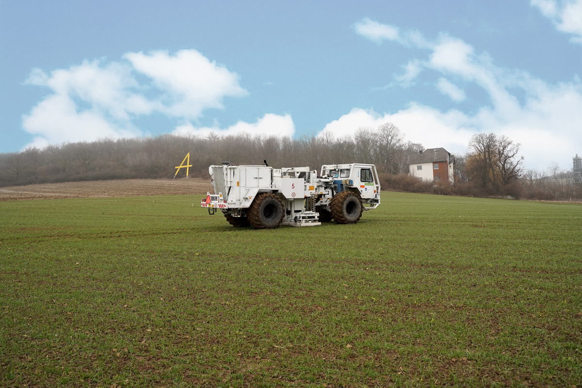 Auf einem Feld steht ein großes weißes Fahrzeug. Im Hintergrund ist ein Wald zu sehen vor blauem Himmel.