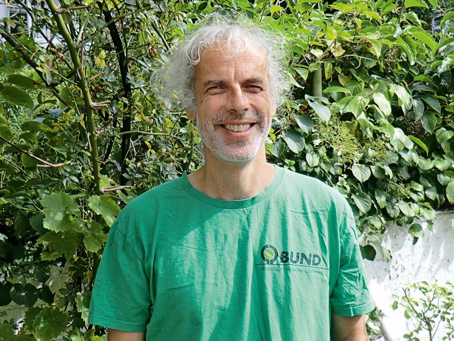 Vor einer Hecke steht ein Mann mit grauen gelockten Haaren und Bart. Er trägt ein grünes T-Shirt und lächelt in die Kamera.