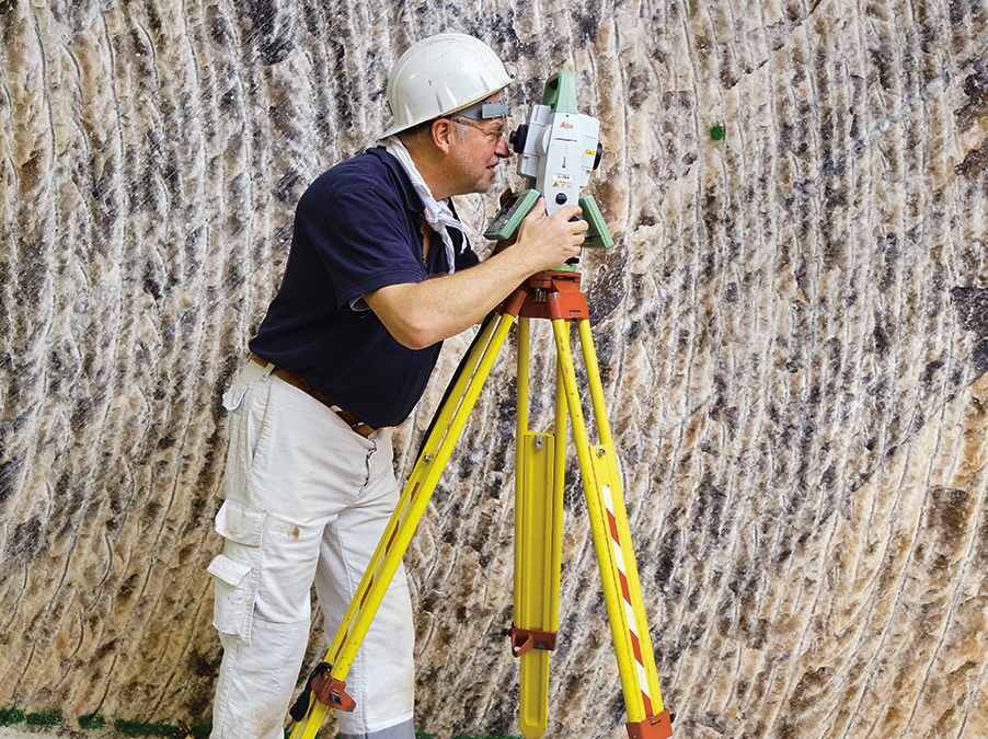 Ein Mann mit weißer Hose und weißem Helm schaut durch ein Nivelliergerät. Er steht leicht gebückt. Im Hintergrund ist eine weiße wellige Wand zu sehen.